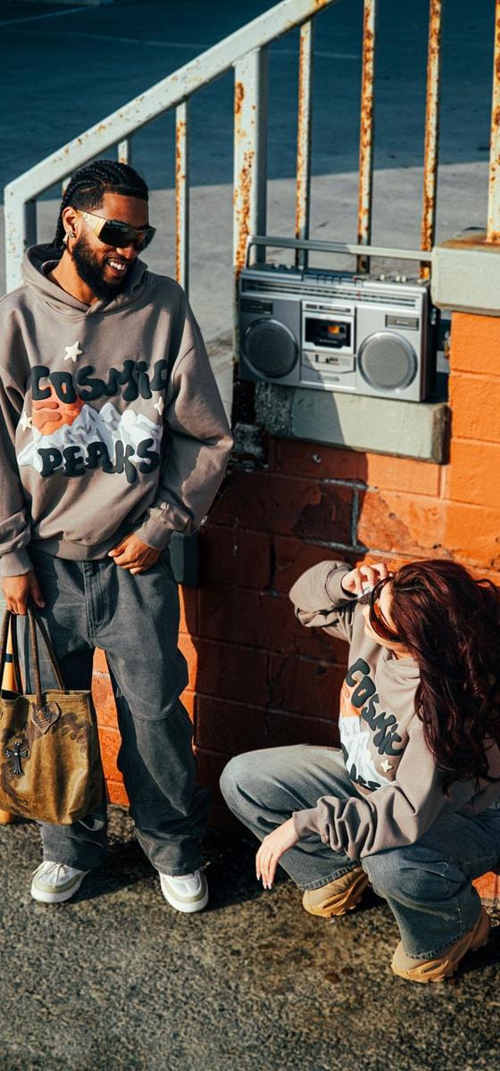 A man and a woman wearing taupe Broken Planet Market Cosmic Peaks hoodies and grey tracksuit bottoms beside an orange brick wall
