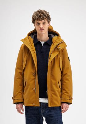 A man wearing an ochre Butcher of Blue hooded parka over a navy knit polo and dark jeans
