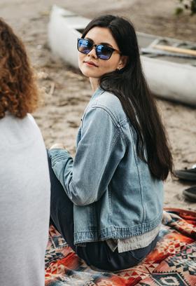 A woman wearing blue-mirrored CAMP Eyewear sunglasses and a denim jacket seated on a blanket by a canoe