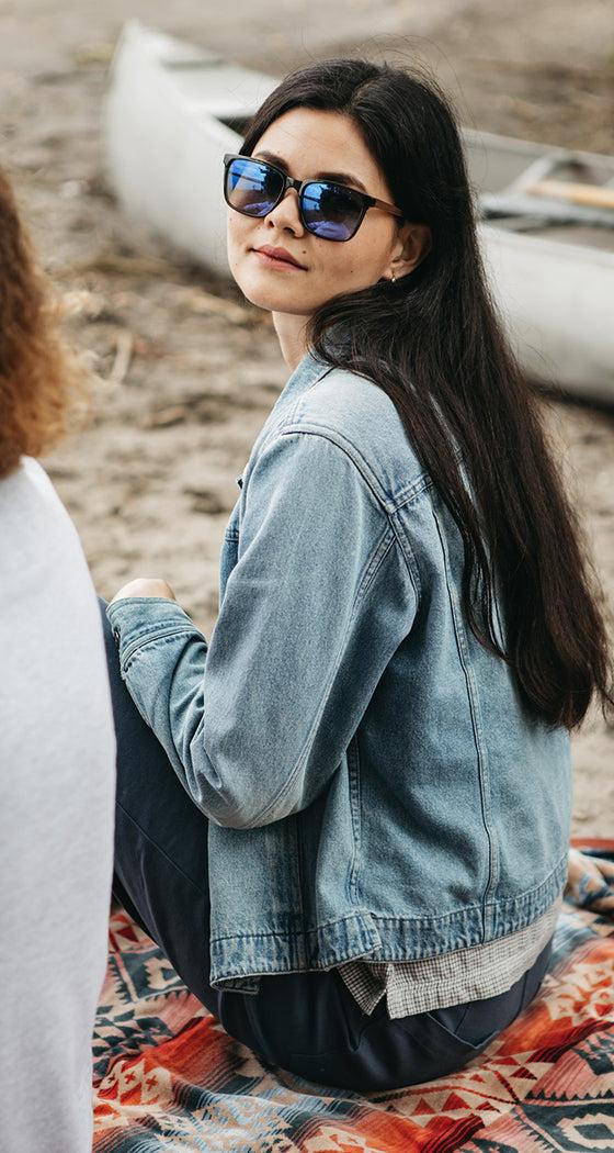 A woman wearing blue-mirrored CAMP Eyewear sunglasses and a denim jacket seated on a blanket by a canoe