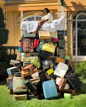 A woman in a white robe reclining on a bed atop a towering stack of vintage suitcases