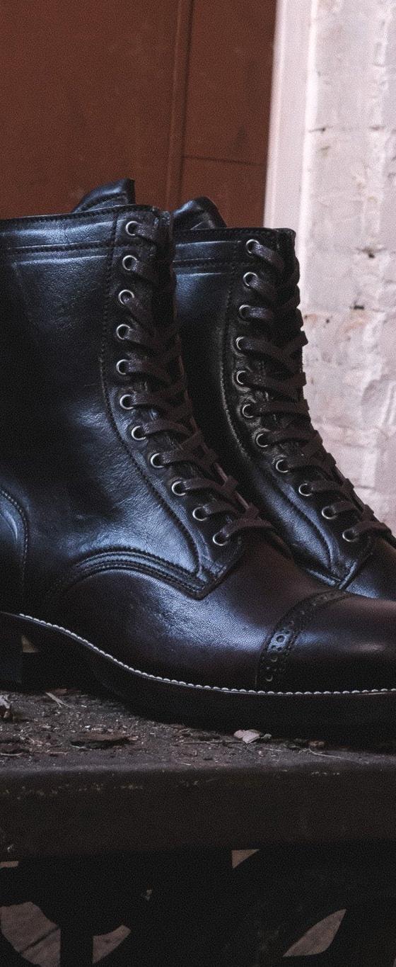 A pair of black cap-toe lace-up leather boots resting on a weathered iron cart against a whitewashed brick wall