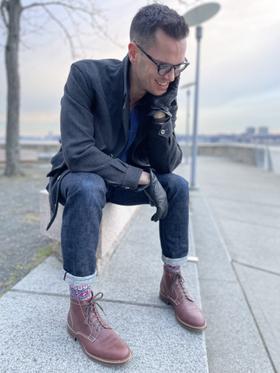 A man in a charcoal overcoat and cuffed selvedge denim seated outdoors, wearing brown Caswell lace-up boots