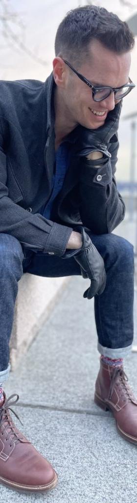 A man in a charcoal overcoat and cuffed selvedge denim seated outdoors, wearing brown Caswell lace-up boots