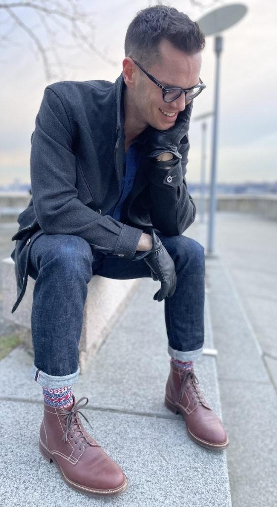 A man in a charcoal overcoat and cuffed selvedge denim seated outdoors, wearing brown Caswell lace-up boots