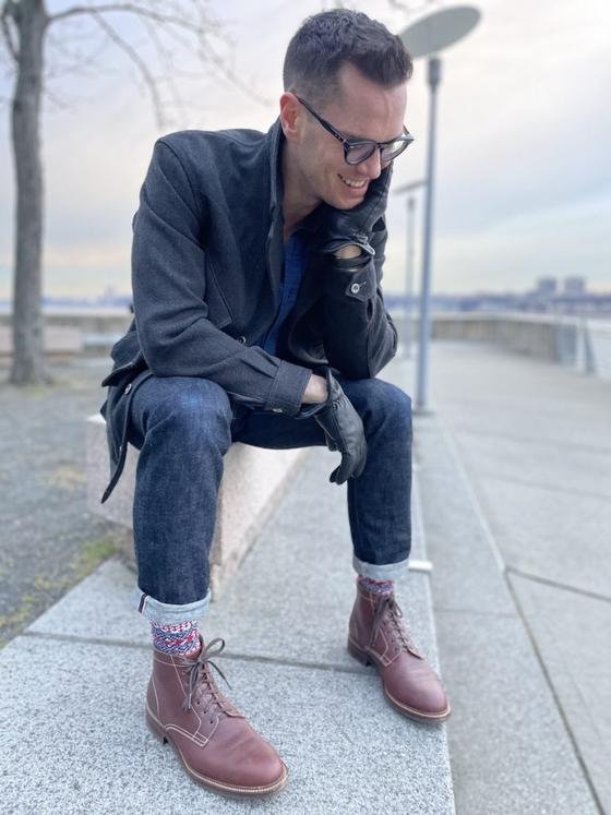 A man in a charcoal overcoat and cuffed selvedge denim seated outdoors, wearing brown Caswell lace-up boots