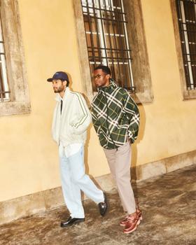 Two men walking outdoors, one in a cream track jacket and light jeans, the other in a green checked overshirt and taupe trousers