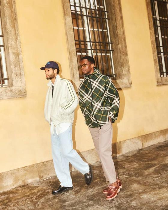 Two men walking outdoors, one in a cream track jacket and light jeans, the other in a green checked overshirt and taupe trousers