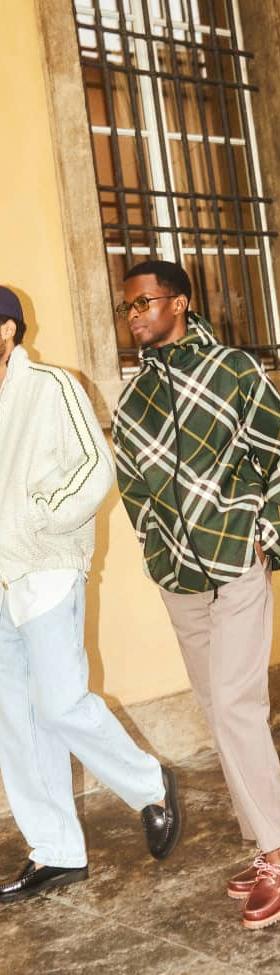 Two men walking outdoors, one in a cream track jacket and light jeans, the other in a green checked overshirt and taupe trousers