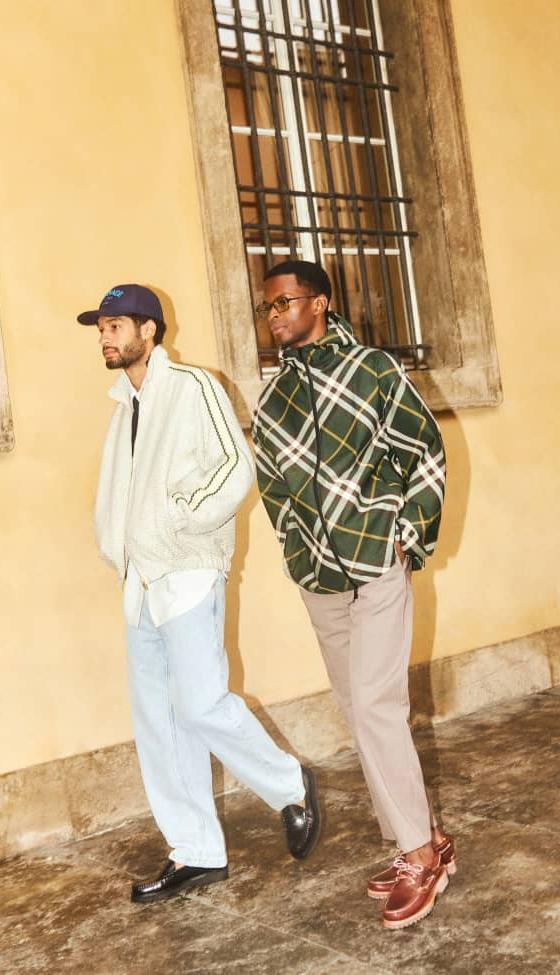 Two men walking outdoors, one in a cream track jacket and light jeans, the other in a green checked overshirt and taupe trousers