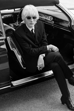 A man in a black skinny Celine Homme suit and sunglasses seated in the doorway of a vintage car