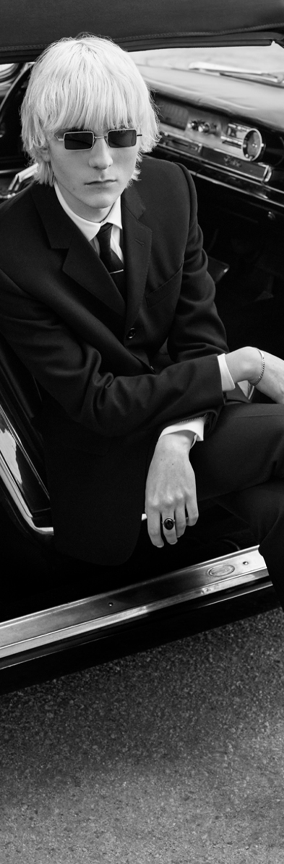 A man in a black skinny Celine Homme suit and sunglasses seated in the doorway of a vintage car