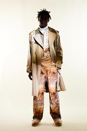 A man wearing a Children of the Discordance layered trench coat with scarf-print patchwork trousers