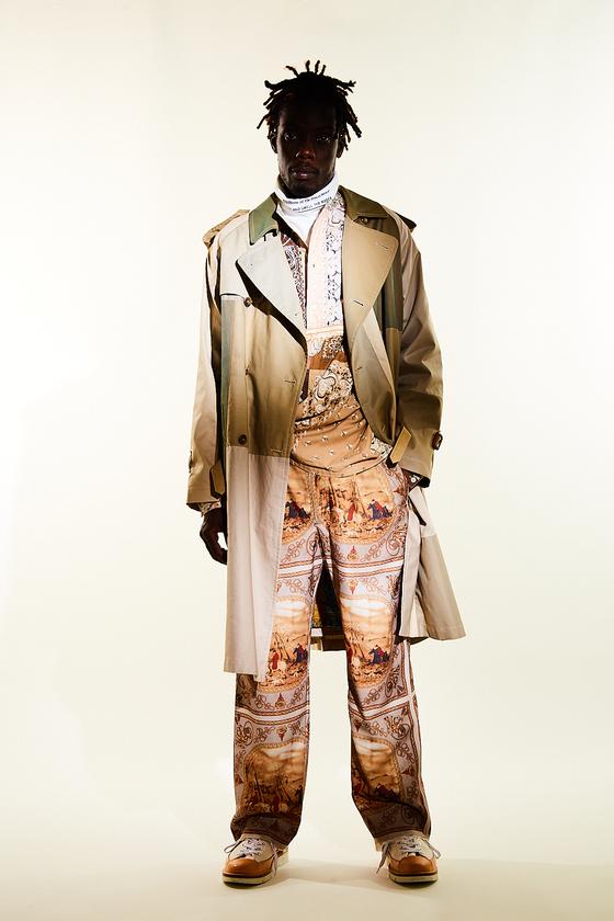 A man wearing a Children of the Discordance layered trench coat with scarf-print patchwork trousers