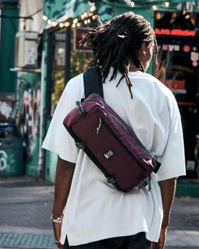 A person wearing a white tee with a burgundy Chrome Industries sling bag across their back on a city street