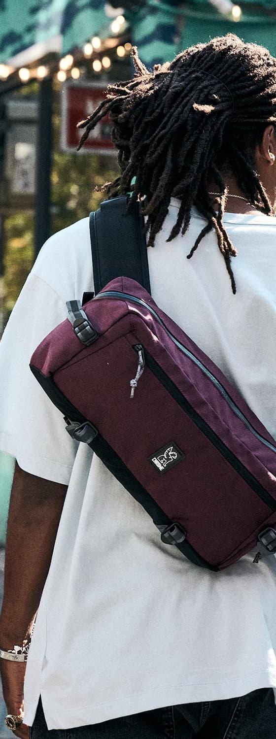 A person wearing a white tee with a burgundy Chrome Industries sling bag across their back on a city street