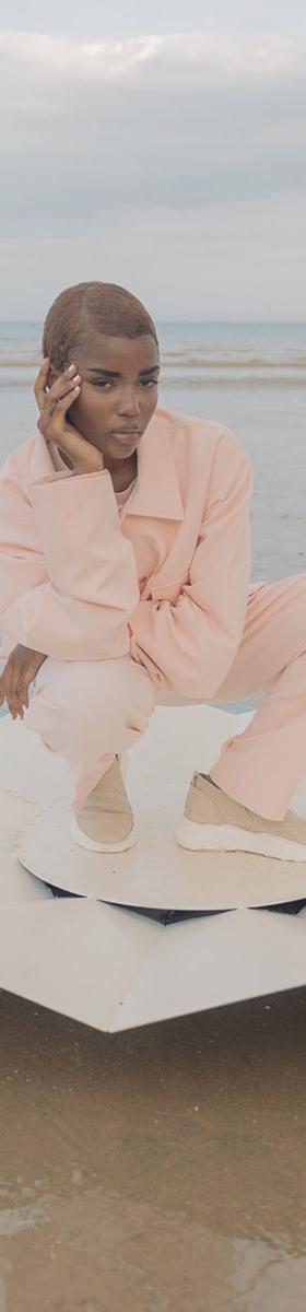 A woman crouching on a beach wearing a pale pink Cold Laundry utility jacket and matching wide-leg trousers