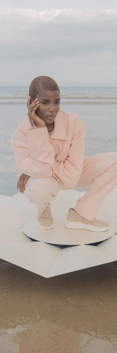 A woman crouching on a beach wearing a pale pink Cold Laundry utility jacket and matching wide-leg trousers