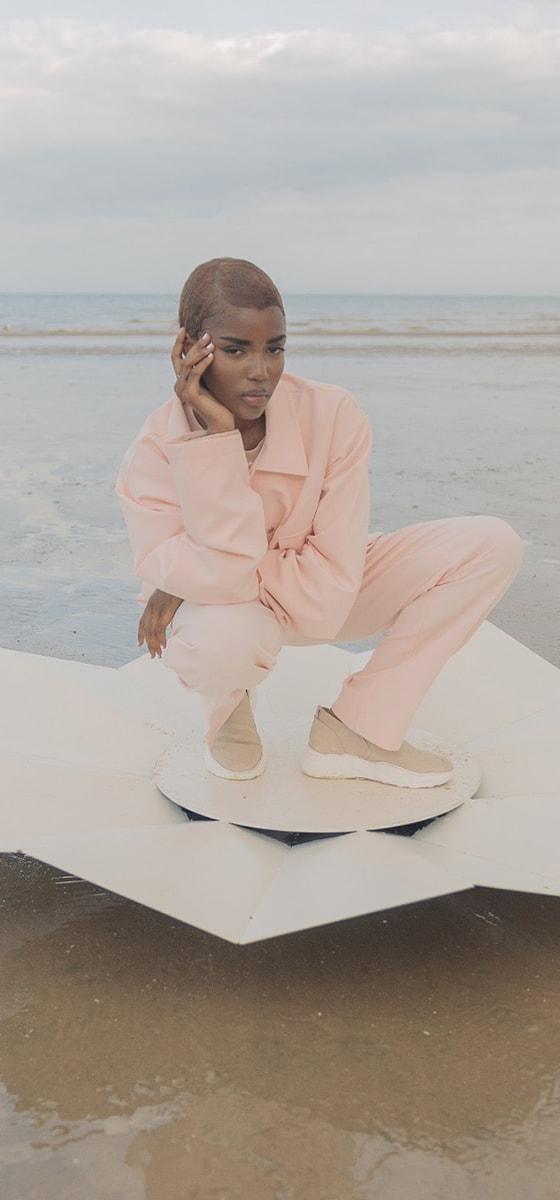 A woman crouching on a beach wearing a pale pink Cold Laundry utility jacket and matching wide-leg trousers