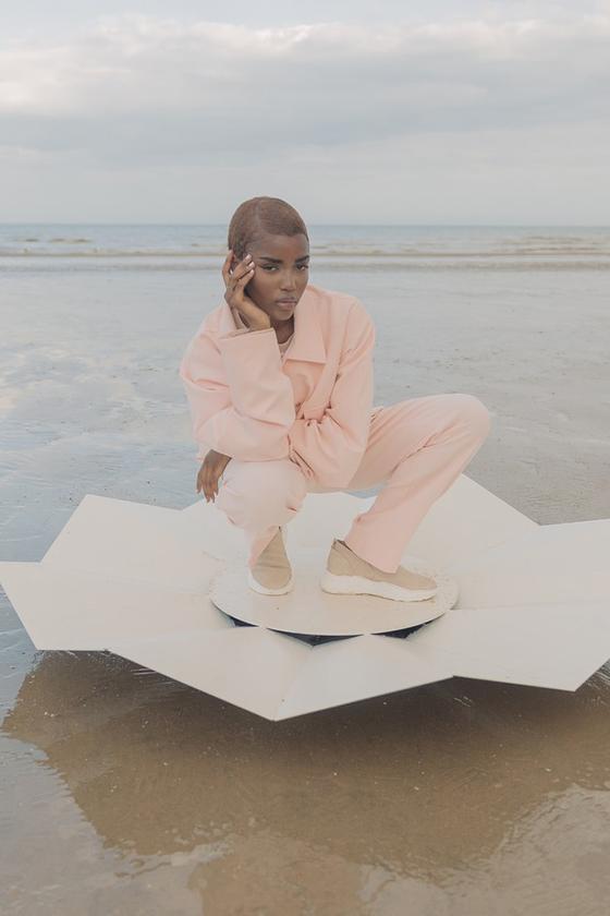 A woman crouching on a beach wearing a pale pink Cold Laundry utility jacket and matching wide-leg trousers