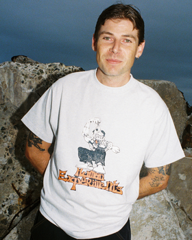 A man wearing a white Come Sundown graphic T-shirt standing against rocks at dusk