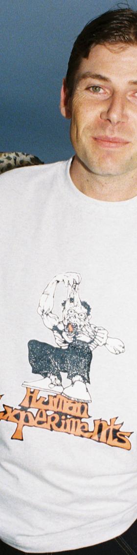 A man wearing a white Come Sundown graphic T-shirt standing against rocks at dusk