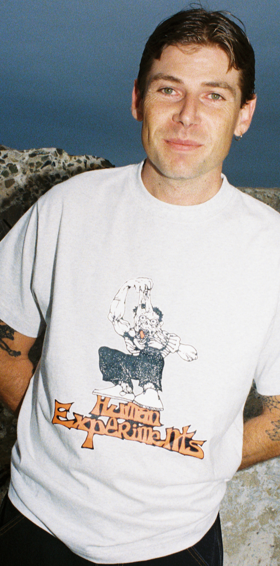 A man wearing a white Come Sundown graphic T-shirt standing against rocks at dusk
