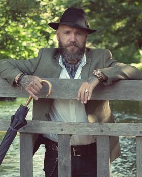 A bearded man in a tweed jacket and felt hat leaning on a wooden bridge railing