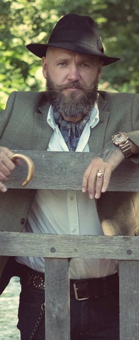 A bearded man in a tweed jacket and felt hat leaning on a wooden bridge railing