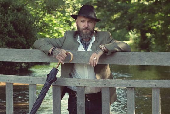 A bearded man in a tweed jacket and felt hat leaning on a wooden bridge railing