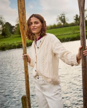 A woman wearing a cream cable-knit Crew Clothing cardigan and white jeans, holding oars beside a river