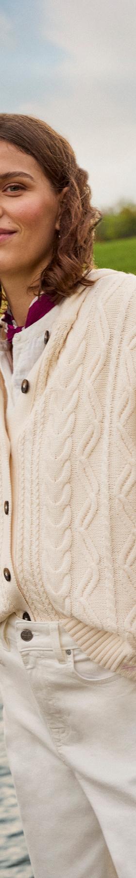 A woman wearing a cream cable-knit Crew Clothing cardigan and white jeans, holding oars beside a river