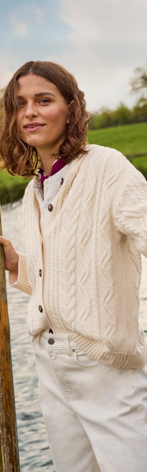 A woman wearing a cream cable-knit Crew Clothing cardigan and white jeans, holding oars beside a river