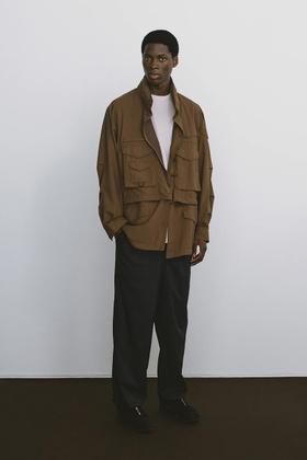 A man wearing a brown DAIWA PIER39 multi-pocket field jacket with black trousers