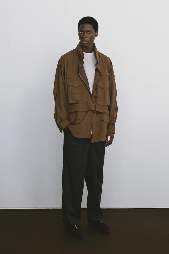 A man wearing a brown DAIWA PIER39 multi-pocket field jacket with black trousers