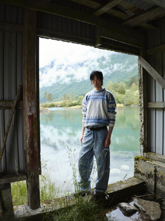 A person wearing a blue and white patterned Dale of Norway wool sweater standing in a boathouse doorway by a fjord