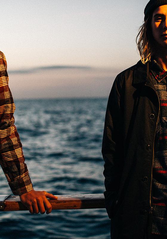 Two men wearing Dark Seas Division plaid flannel shirts and a dark work jacket at sea