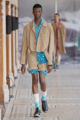 A man wearing a cropped tan denzilpatrick jacket over a turquoise shirt and printed shorts