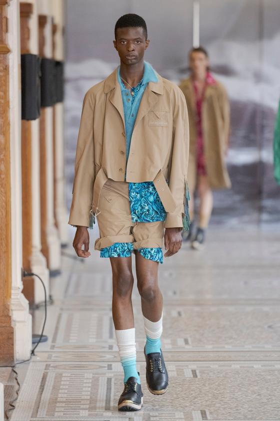 A man wearing a cropped tan denzilpatrick jacket over a turquoise shirt and printed shorts