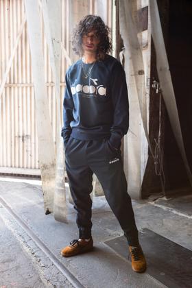 A man wearing a navy Diadora sweatshirt and matching track pants with tan suede sneakers