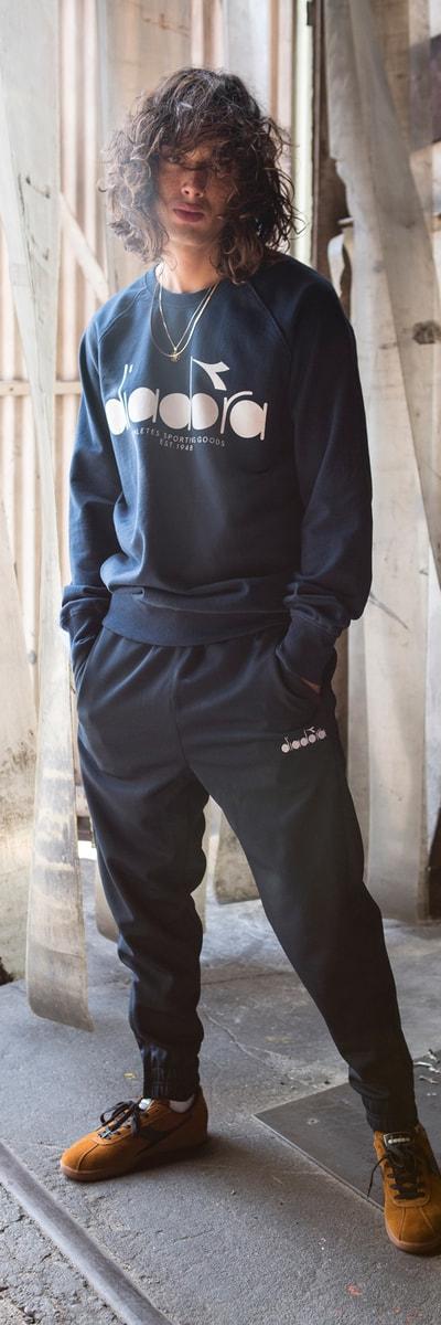 A man wearing a navy Diadora sweatshirt and matching track pants with tan suede sneakers