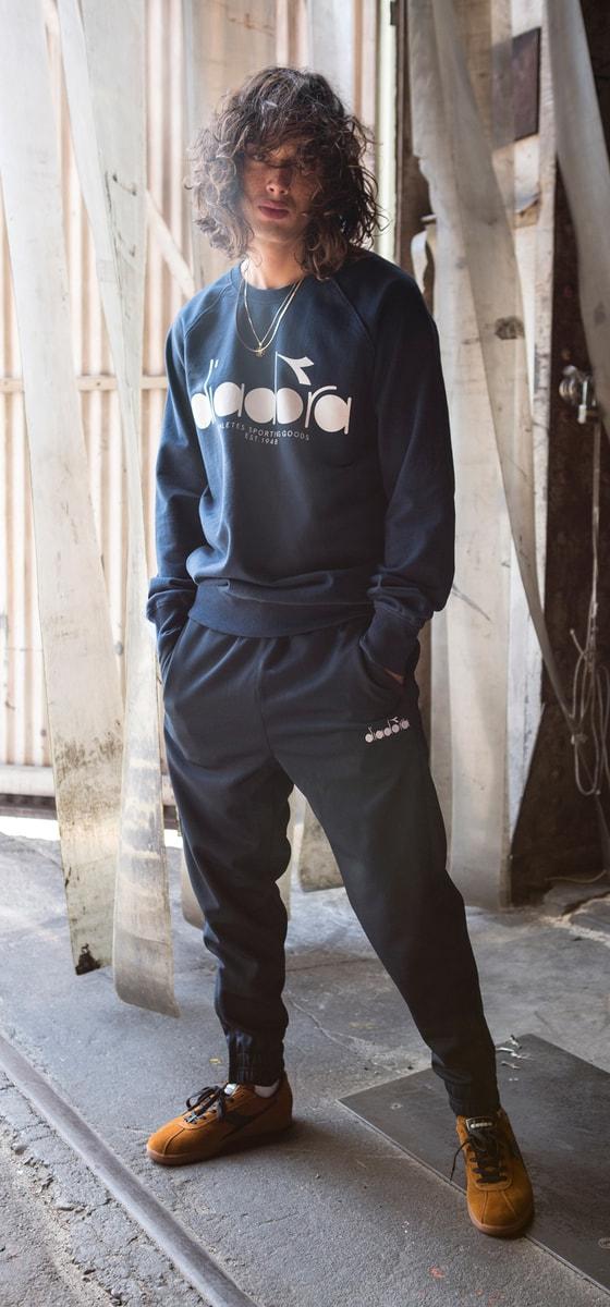 A man wearing a navy Diadora sweatshirt and matching track pants with tan suede sneakers