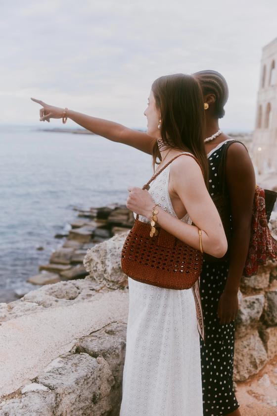 A woman in a white eyelet dress carrying a woven tan leather Dragon Diffusion shoulder bag by the sea