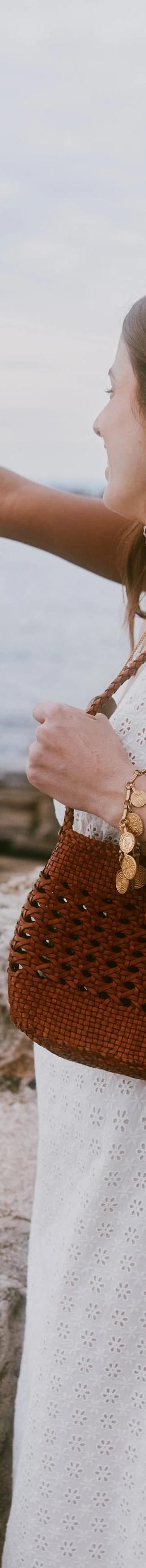 A woman in a white eyelet dress carrying a woven tan leather Dragon Diffusion shoulder bag by the sea