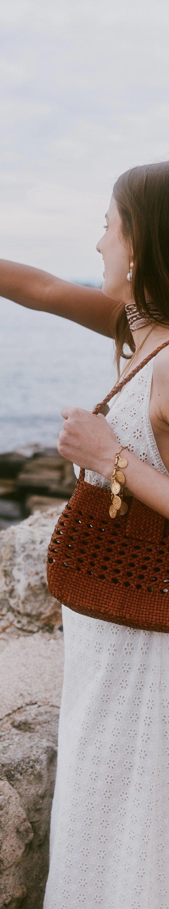 A woman in a white eyelet dress carrying a woven tan leather Dragon Diffusion shoulder bag by the sea