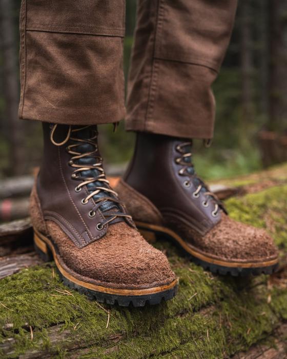 A man wearing brown roughout leather Drew's logger boots and waxed canvas trousers, standing on a mossy log in an Oregon forest