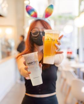A woman in a black top holding two Drip Tea drinks in a bright cafe