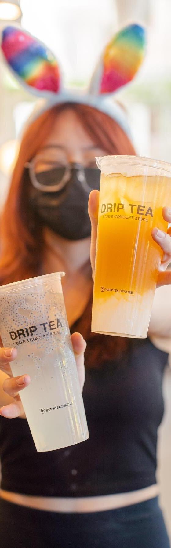 A woman in a black top holding two Drip Tea drinks in a bright cafe