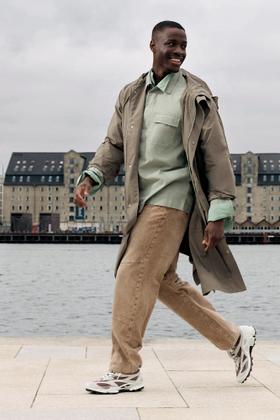 A man walking along a Copenhagen waterfront in a taupe trench coat, sage overshirt and chunky white Ecco sneakers