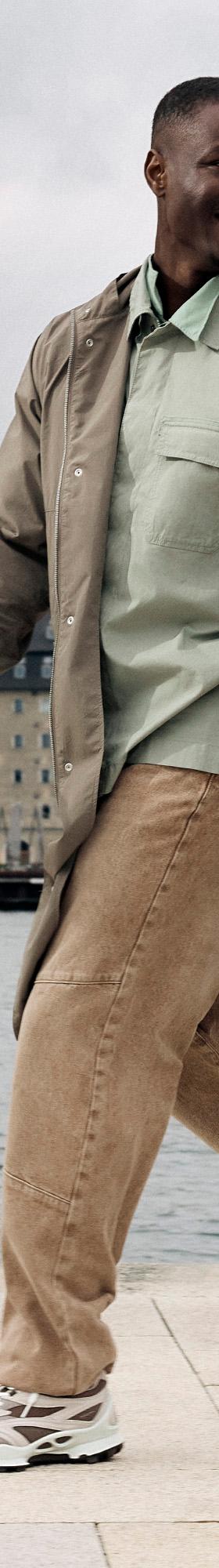 A man walking along a Copenhagen waterfront in a taupe trench coat, sage overshirt and chunky white Ecco sneakers
